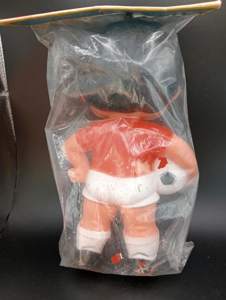 Oude voetbalspelerfiguur van hard plastic met rood shirt, witte korte broek, zwart-witte bal en transparant zakje met blauwe kartonnen kop