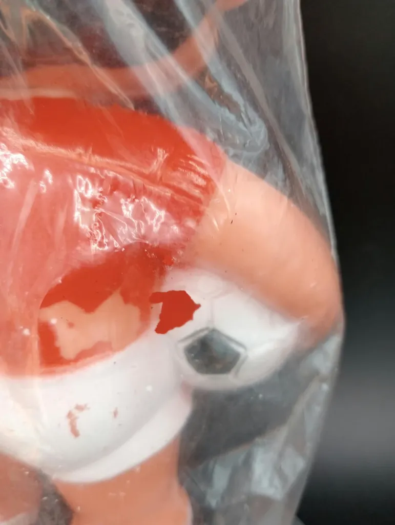 Alte Fußballspielerfigur aus Hartplastik mit rotem Trikot, weißer Hose, schwarz-weißem Ball und transparentem Beutel mit blauem Kartonkopf