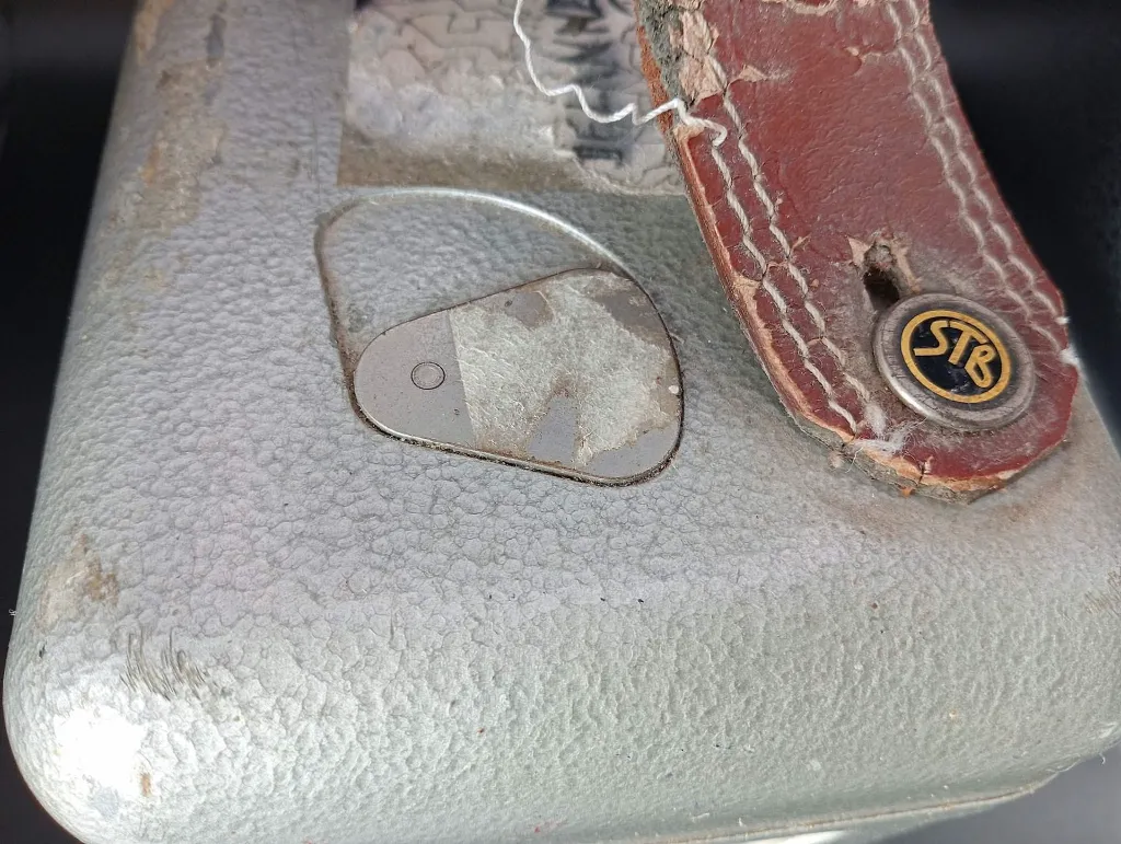 Oude mechanische STB Swiss Made duivenklok van grijs gehamerd metaal met witte wijzerplaat nummer 67992, zijdelingse sluiting met metalen verzegeling en sterk gebarsten rood leren handvat