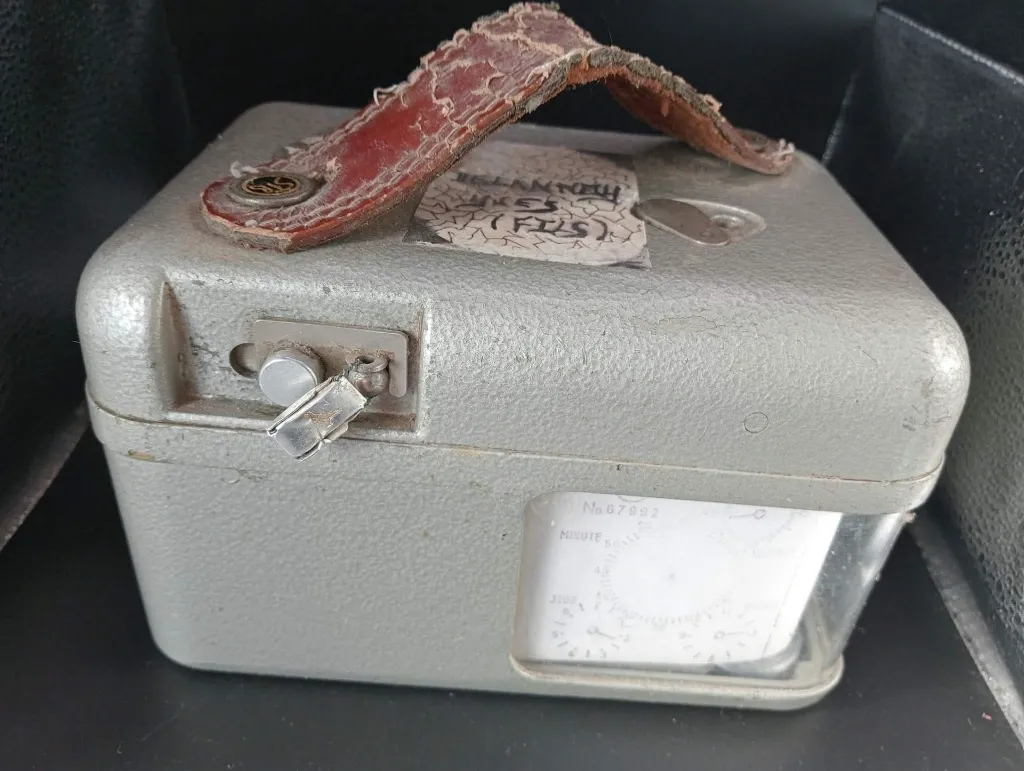 Antigo relógio colombófilo STB Swiss Made em metal cinzento martelado com mostrador branco número 67992, fecho lateral com selo metálico e pega vermelha muito gretada