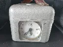 Ancien constateur colombophile mécanique STB Swiss Made en métal gris martelé avec cadran blanc numéro 67992, fermeture latérale munie d’un scellé métallique et poignée rouge en cuir fortement craquelée