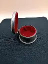 Caixa oval metálica prateada com interior de veludo vermelho.